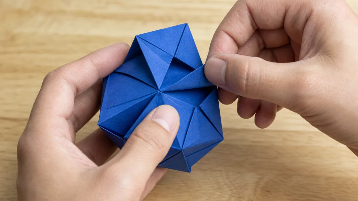 hands carefully folding a complex geometric origami model from deep blue paper…