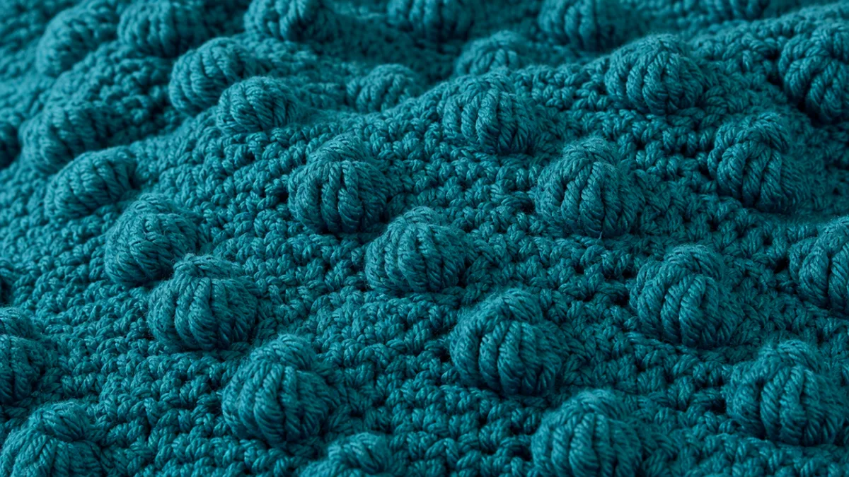close-up of a rich teal crochet blanket featuring raised popcorn stitch clusters,…
