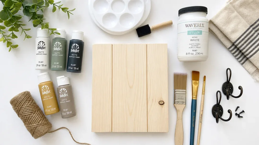a flat lay of DIY crafting materials including paints, brushes, wooden panel,…