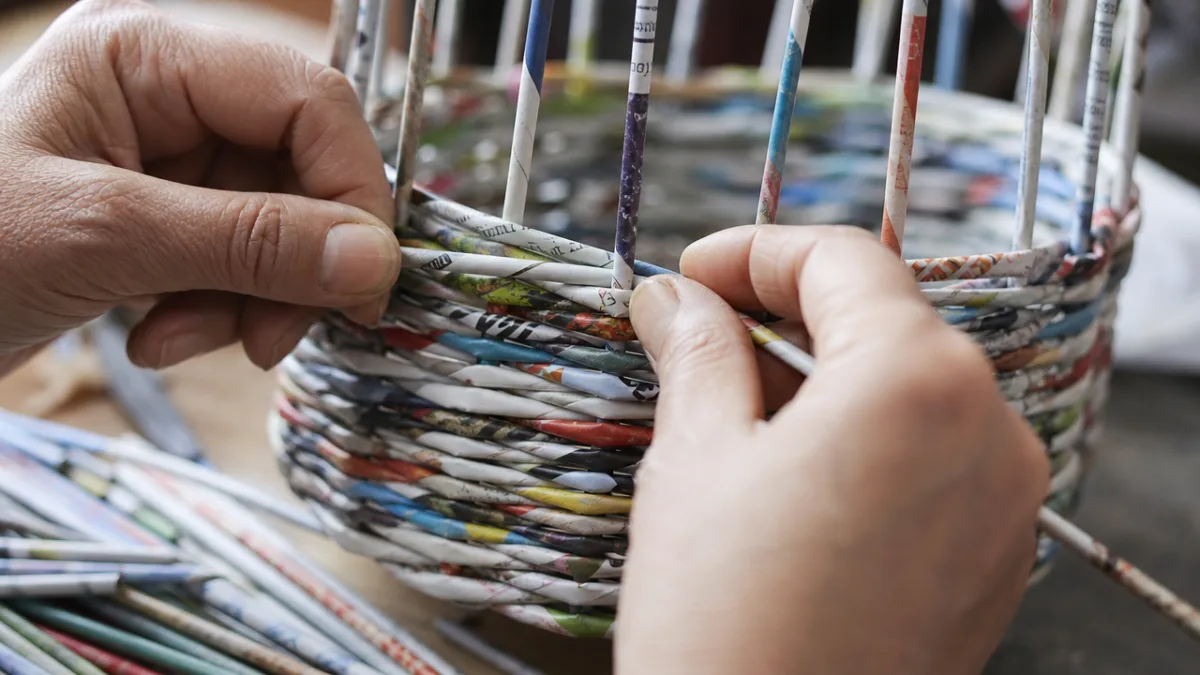 hands weaving colorful rolled magazine tubes into a basket shape, close-up shot…