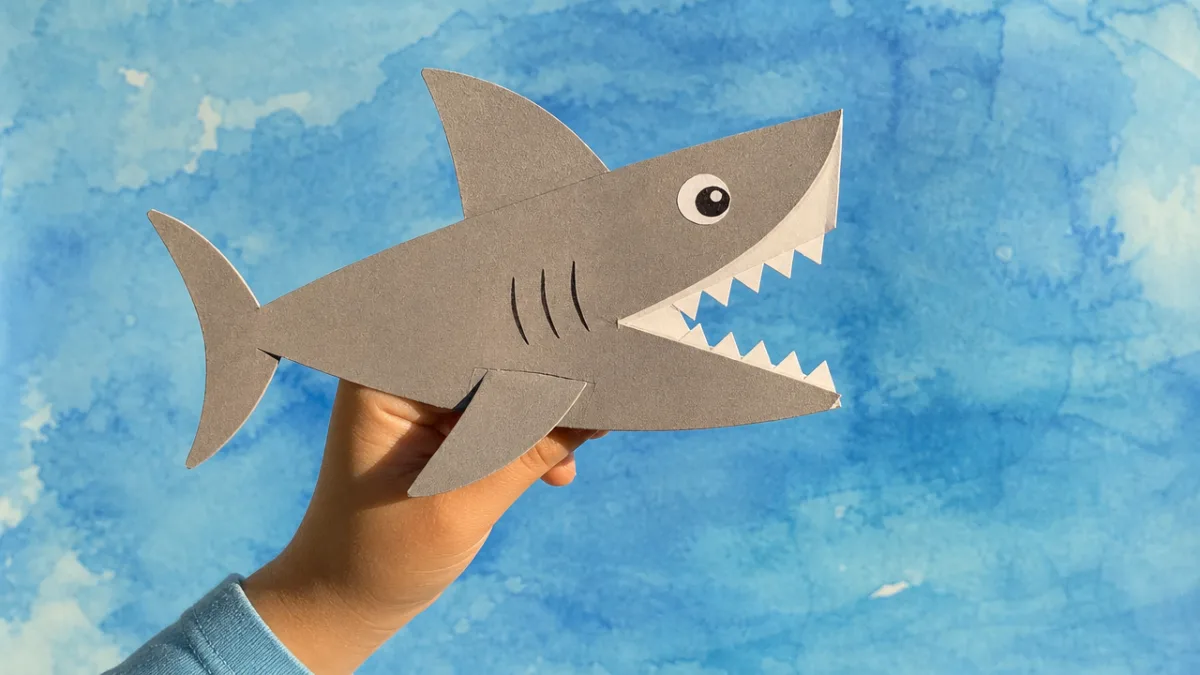 a finished grey shark paper puppet held up against a blue watercolor…