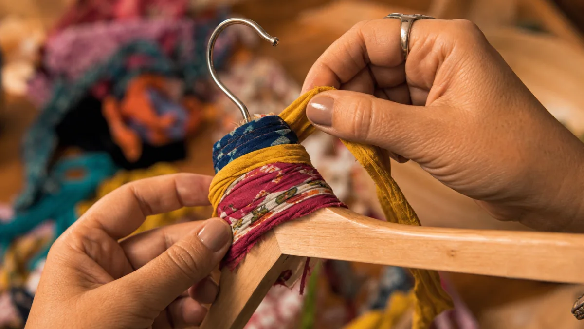 close-up of hands wrapping colorful fabric strips around a wooden hanger, warm…