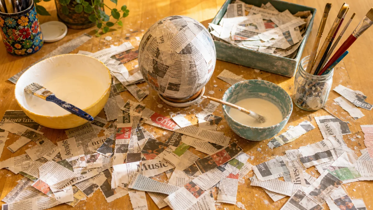 a bright craft table covered in torn newspaper strips with bowls of…
