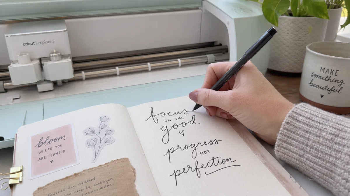 a close-up of a crafter's hand writing sayings in a journal beside…